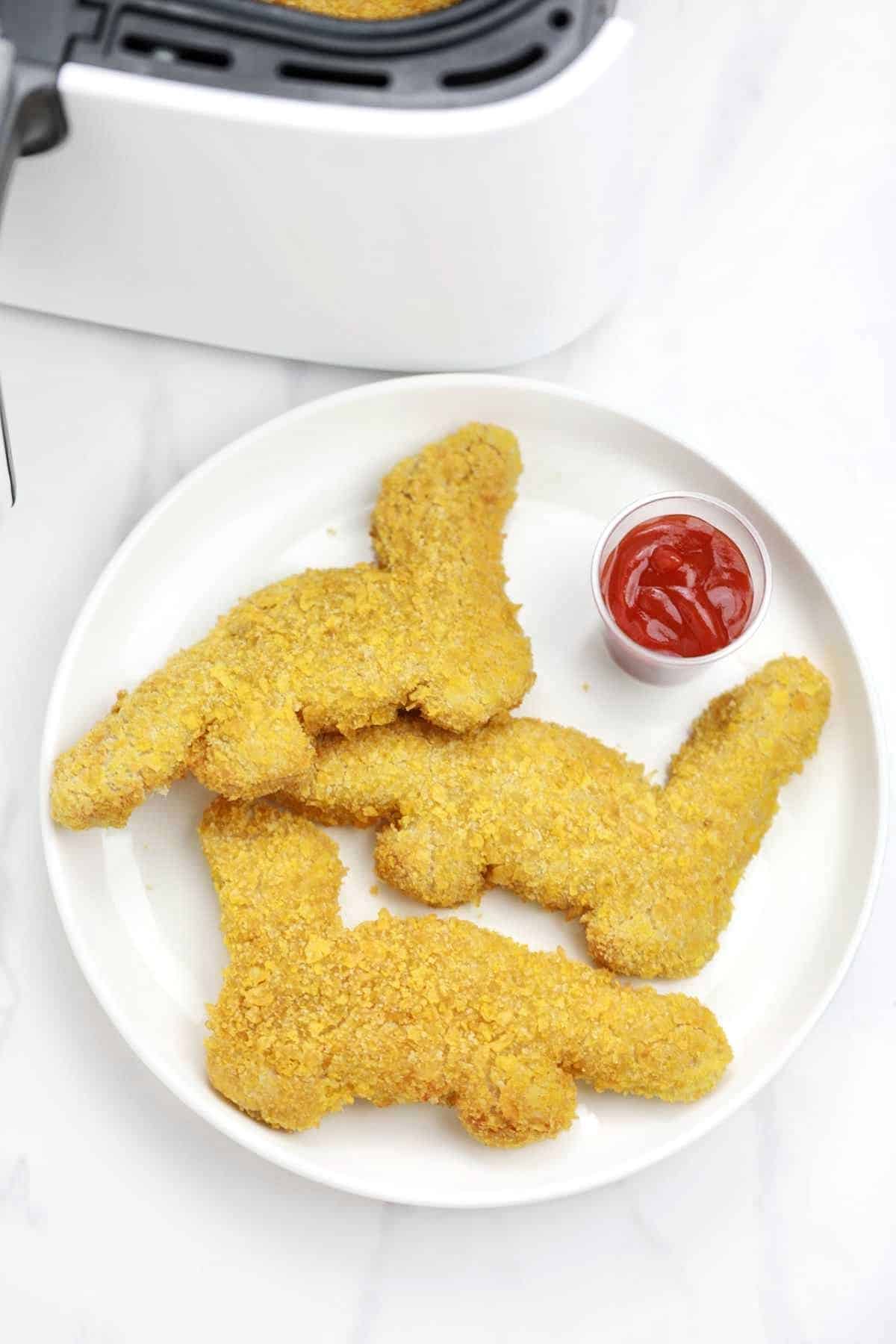 3 pieces dinosaur nuggets served on a plate with  ketchup.